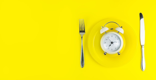 Будильник с вилкой и ножом на светло-желтом фоне. время поесть, завтрак, обед и ужин концепции. копировать пространство, баннер