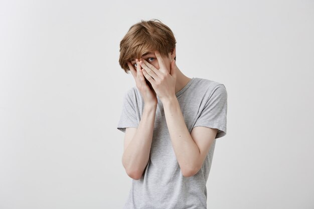 Alarmed nervous caucasian guy covers face with hands, being shocked or surprised to see unexpected gift prepared by girlfriend. Astonished emotional fair-haired young male against gray background