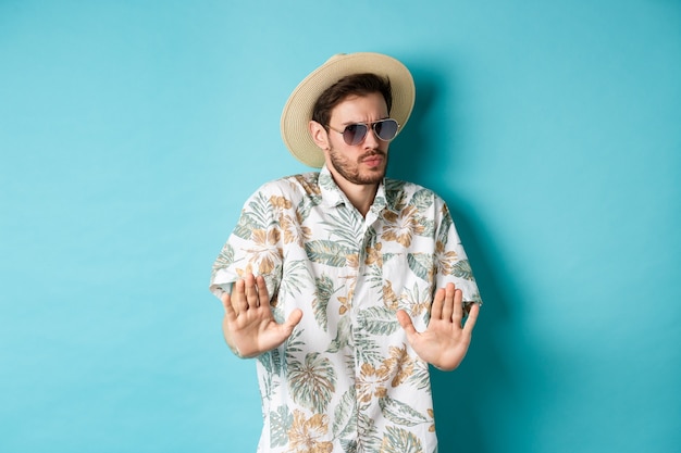 Встревоженный турист просит держаться подальше, отойти от чего-то съеживается, показывая жест отказа, стоит в соломенной шляпе и гавайской рубашке, синий фон.