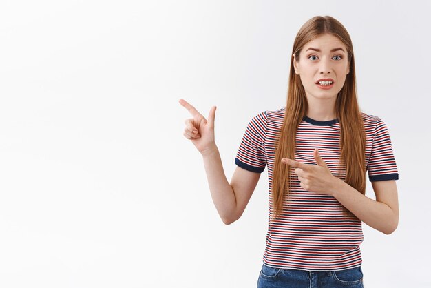 Alarmed and worried young girl with long hair feeling anxious about doubtful strange action happening pointing fingers left squinting and cringe bothered standing worried and embarrassed