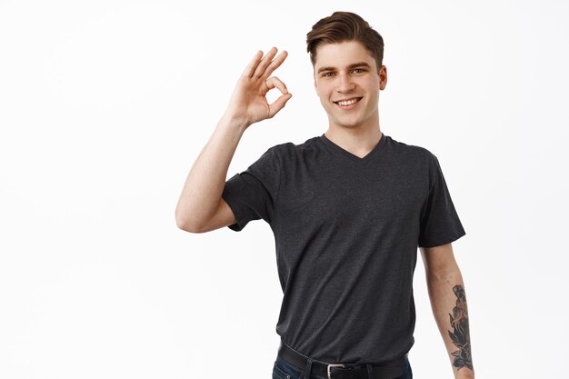 All under control. Smiling young man say okay, show zero OK gesture and nod in approval, recommend something good, likes and approves brand product, white background