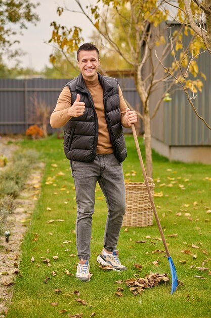모두 훌륭합니다. 가을날 정원에 있는 녹색 잔디밭에 갈퀴를 들고 갈퀴를 들고 서 있는 스웨터 조끼와 청바지를 입은 즐거운 매력적인 젊은 성인