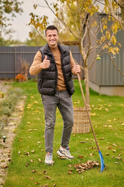 All excellent. Joyful attractive young adult man in sweater vest and jeans standing with rake showing ok gesture on green lawn in garden on autumn day