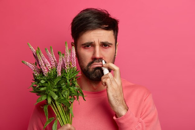 Аллергический бородатый мужчина держит растение, использует капли в нос для лечения чихания, у него недовольное выражение лица, реакция на аллерген, излечивает морскую болезнь, следует советам аллерголога. Концепция здравоохранения