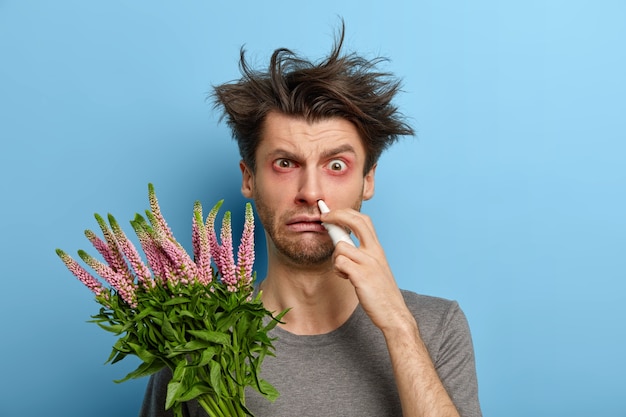 Allergic man has messy hairstyle, red itchy eyes, holds plant causing sneezing or stiff, suffers from unpleasant symptoms, has home treatment, stands against blue wall. Nasal discharge