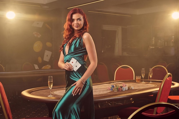 Alluring redheaded girl in a long blue satin dress is smiling and posing sideways with two aces in her hand against a poker table in luxury casino. Passion, cards, chips, alcohol, win, gambling - it i