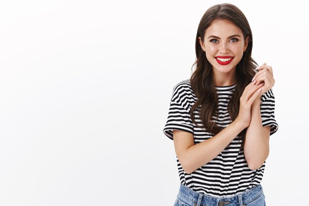 Alluring tender young female in red lipstick, striped t-shirt, rubbing hands together gently touching arms, smiling joyfully, happily stand white wall