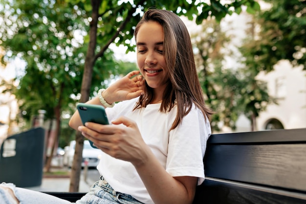 Alluring woman with loose hair in white short is touching her hair and looking at smartphone while sitting on city bench and waiting for friends