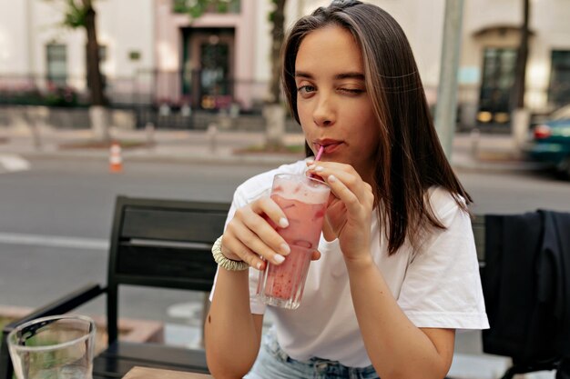 Alluring young woman winking at camera and smiling while drinking summer smoothie and sitting on terrace closeup The bare and thin curves of collarbones are striking