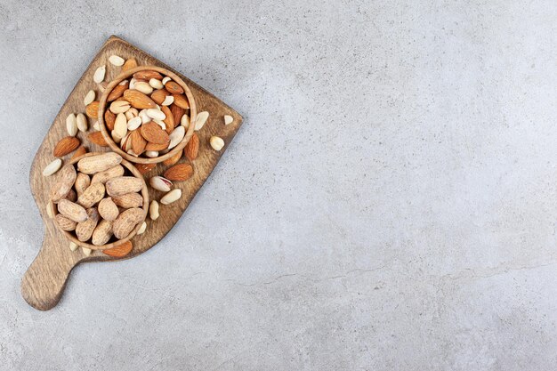 Almonds, pistachios and peanuts heaped in and around wooden bowls on a wooden board on marble surface