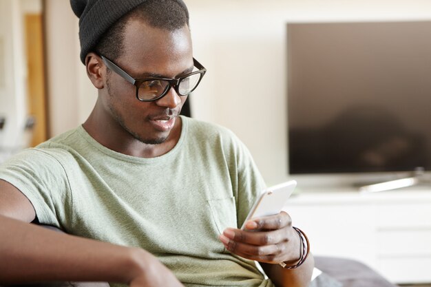 Always in touch. Cheerful young dark-skinned man wearing hipster hat and rectangular glasses browsing social media on electronic device at home