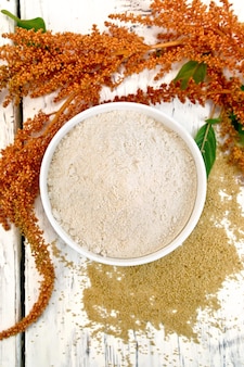 Amaranth flour in a bowl, seeds scattered on the table, brown flower with green leaves on the background of the wooden planks on top
