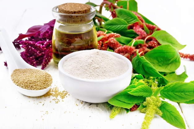 Amaranth flour in a bowl, seeds in a spoon and oil in a glass jar, brown, green and purple flowers of a plant on wooden board background