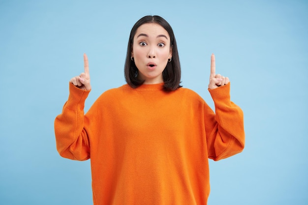 Amazed asian female model says wow looks impressed points fingers up stands over blue background