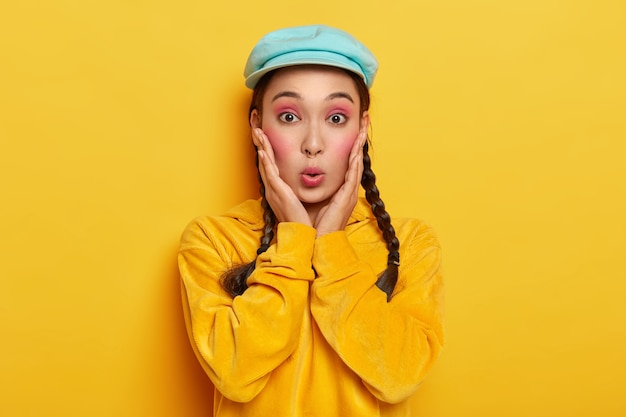 Amazed Asian lady keeps lips rounded, looks with surprise, reacts on gossip, wears stylish hat and corduroy yellow sweatshirt