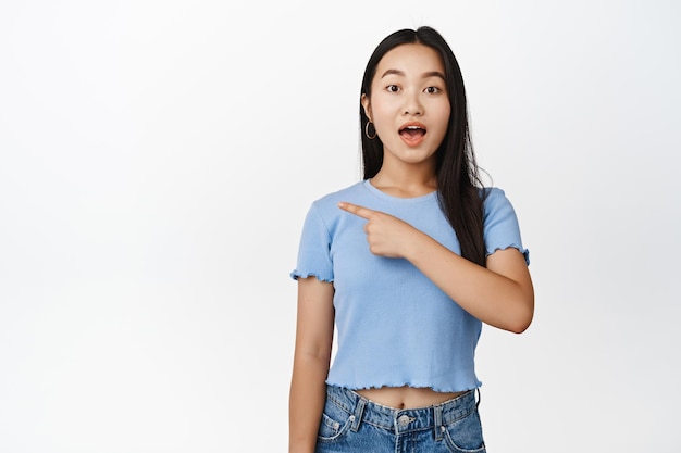 Amazed asian teen girl pointing left gasping and looking excited enthusiastic about promo offer standing over white background