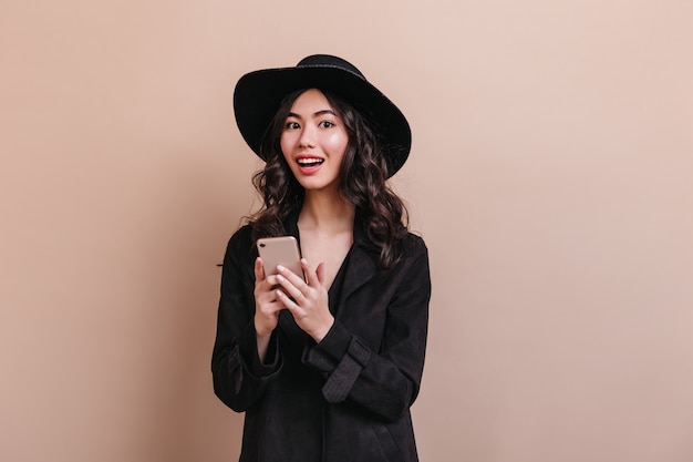 Amazed asian woman holding smartphone and looking at camera. Elegant curly woman in coat posing with gadget.