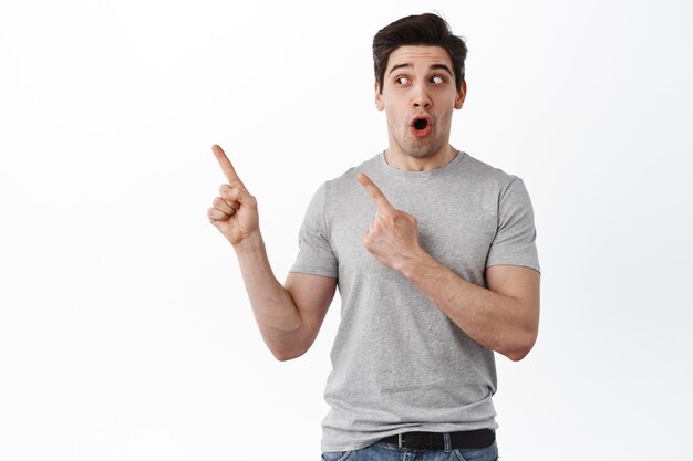 Amazed attractive man points aside, gasps and say wow with excited face, looking at awesome promo deal, standing over white background