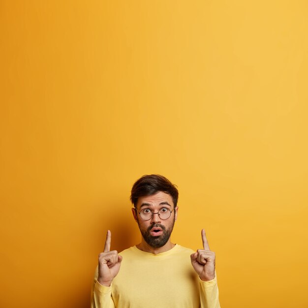 Amazed bearded male student points above with fore fingers, demonstrates new product, discusses sales, gasps from fear, poses over yellow wall, blank space for your promotional content.