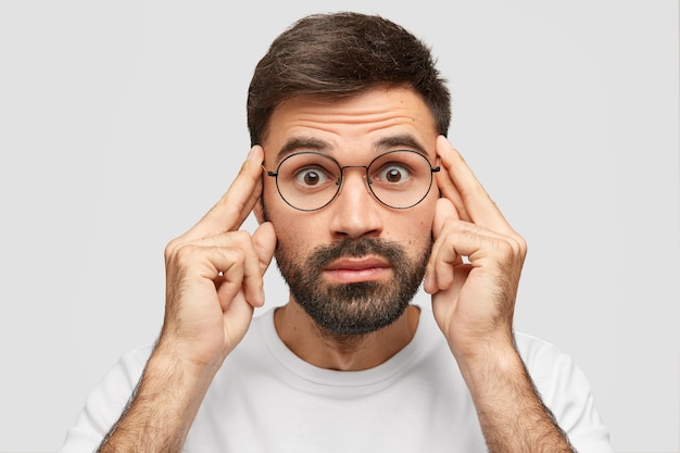 Amazed bearded man keeps fore fingers on temples, looks with stupefied expression, tries to remember details of robbery