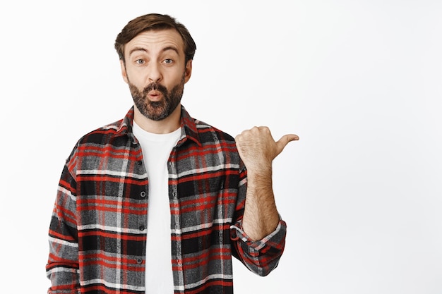 Amazed bearded man say wow looking at camera astonished and pointing right at promo standing in checked shirt over white background