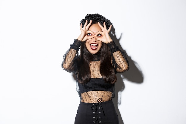 Amazed beautiful asian woman in halloween costume looking through finger glasses with excited happy face, standing over white background wearing gothic witch dress.