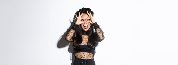 Amazed beautiful asian woman in halloween costume looking through finger glasses with excited happy