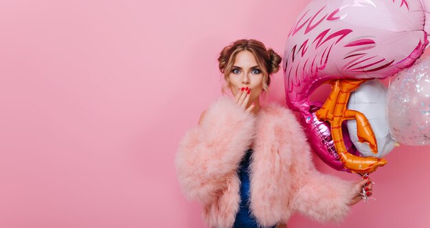 Пораженная красивая девушка в модной шубе прикрывает рот рукой, стоя изолированной на розовом фоне. Удивленная молодая женщина в пушистой куртке держит воздушные шары и ждет друзей