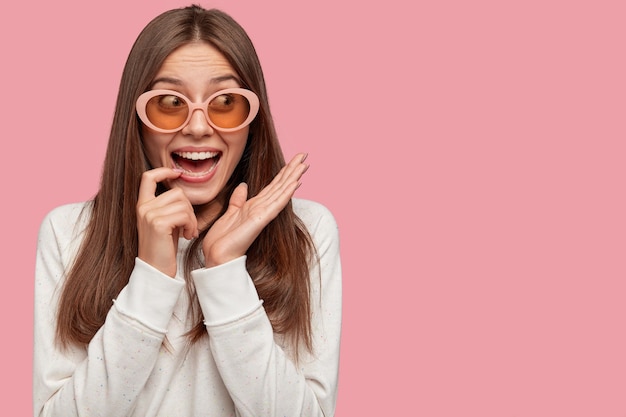 Bella giovane donna stupita con i capelli scuri, indossa grandi sfumature alla moda, sorride ampiamente alla macchina fotografica