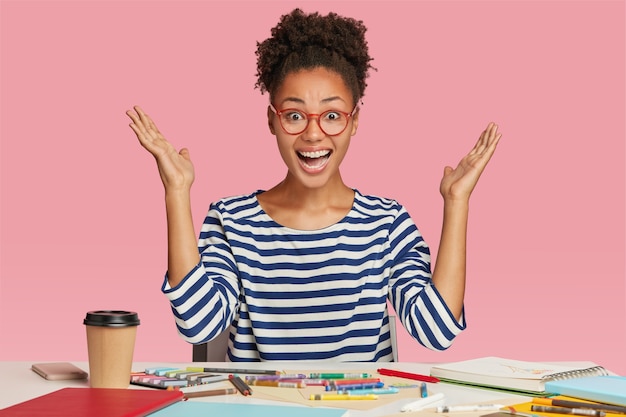 Free photo amazed black female illustrator raises hands in eureka gesture, wears striped clothes