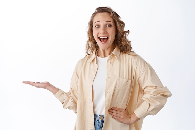 Amazed blond girl gasping and say wow, holding your logo or product in hand, display on palm, standing over white wall