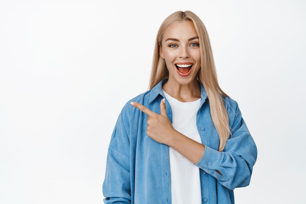 Amazed blond girl points finger left and looks impressed, shows advertisement, stands on white