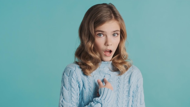 自分自身に指を指して、青い背景の上にカメラでショックを受けているように見えるセーターに身を包んだ驚いた金髪の10代の少女