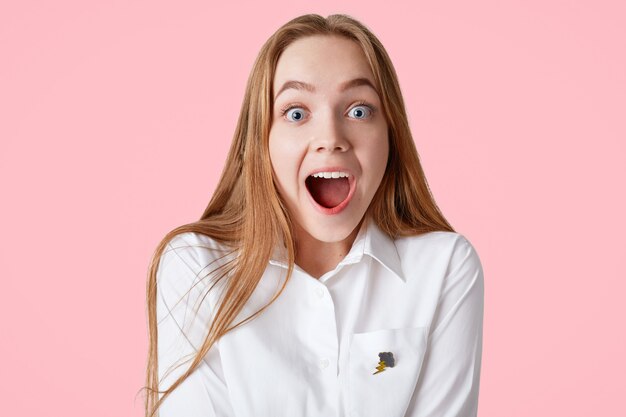 amazed blue eyed female with shocked excited expression, keeps mouth wide opened, recieves unexpected news, isolated on pink wall. People, emotions, reaction concept