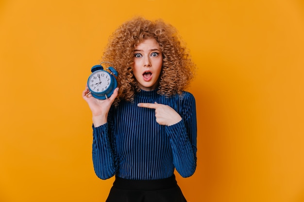 Amazed blue-eyed woman with curls wearing blue top shows alarm clock on yellow space.