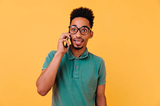 Amazed boy in big glasses talking on phone. Indoor portrait of emotional african guy in green t-shirt calling someone.