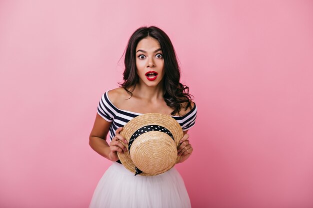 Amazed brown-haired woman looking with mouth open. Spectacular caucasian girl with straw hat in hands standing.