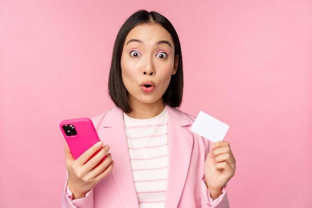 ピンクの背景の上に立っているスマートフォンでクレジットカードと携帯電話の注文オンラインショッピングを示すスーツの驚いた実業家アジアの女の子広告コンセプト
