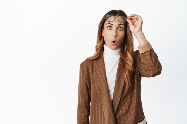 Amazed businesswoman takes off glasses and saying wow impressed by something standing in suit over white background