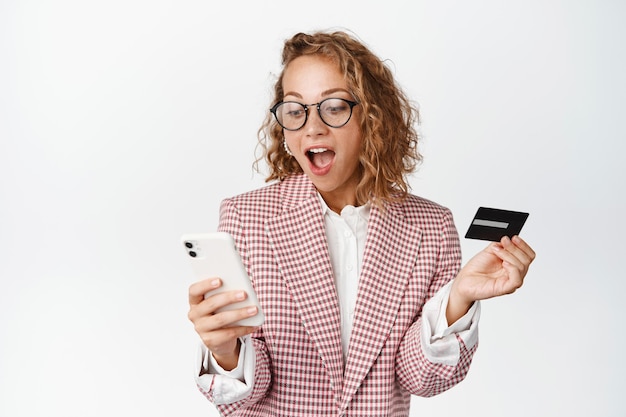 Изумленная корпоративная женщина в костюме смотрит на свой мобильный телефон, держит кредитную карту, платит смартфоном, заказывает онлайн, стоя на белом фоне