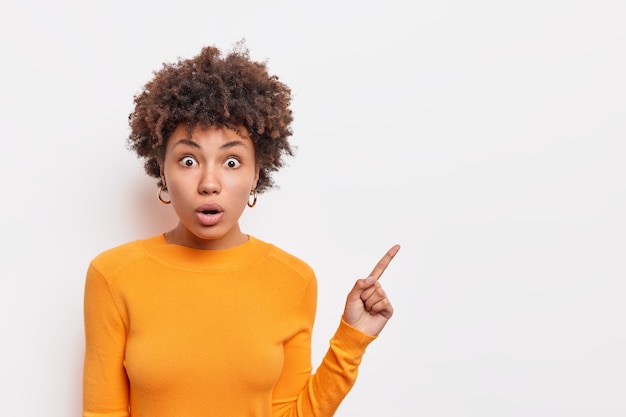 Amazed curly haired young woman with shocked eexpression has stunned face expression wears orange jumper points aside on blank space isolated over white wall. Copy space for advertisement