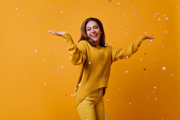 Amazed cute girl with dark hair dancing on yellow wall.  appealing stylish woman throwing out confetti.