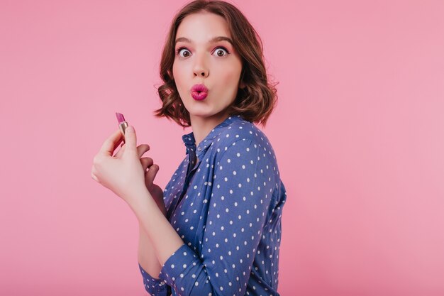 Amazed cute lady with trendy hairstyle doing her makeup on bright pink wall. glamorous girl in stylish blouse holding lipstick.