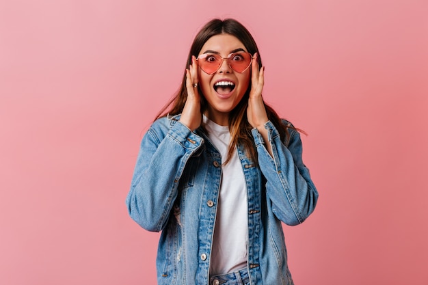 Amazed cute woman looking at camera with open mouth. Front view of surprised girl in denim jacket isolated on pink background.