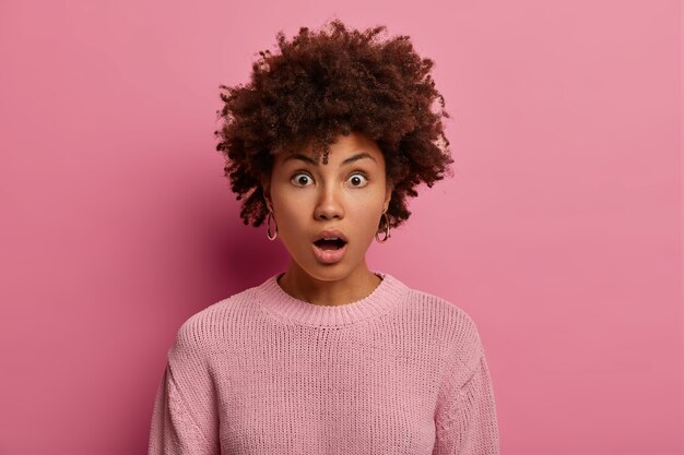 Free photo amazed dark skinned woman gasps from wonder, opens mouth with excitement, learns details and finds out truth of horrible event, wears pink jumper, poses indoor, hears rumors, isolated on rosy wall