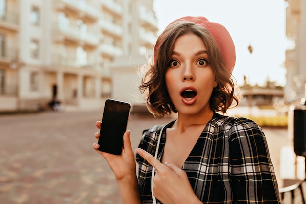 Amazed debonair woman holding smartphone on street. Outdoor photo of shocked emotional girl in red beret.