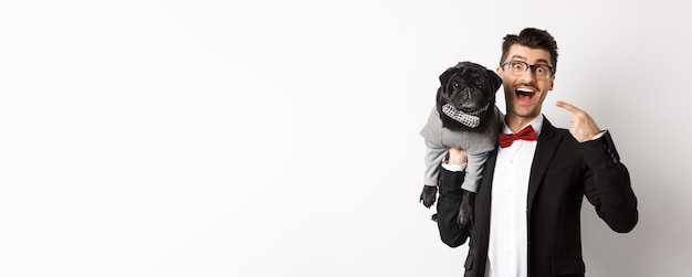 Бесплатное фото Изумленный владелец собаки указывает на своего милого черного мопса, улыбающегося счастливого щенка в костюме на белом фоне