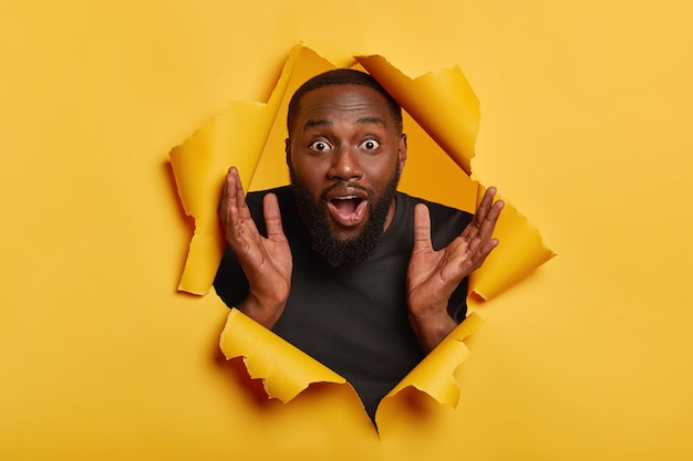 Amazed emotional Afro American man raises hands up, stares with surprised expression, gasps from wonder, being unshaven, clasps palms, poses through hole in yellow paper