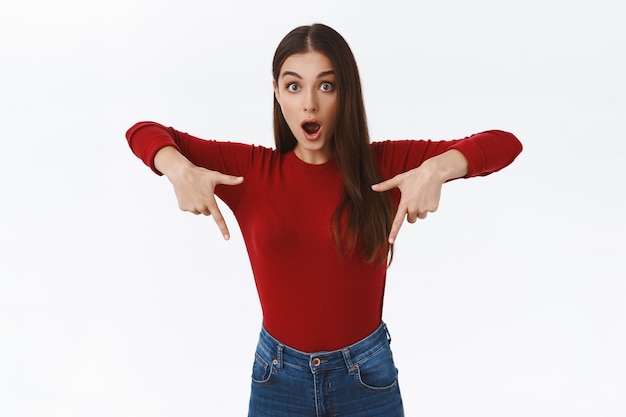 Amazed, enthusiastic speechless pretty caucasian woman in red sweater, drop jaw from amazing cool promo, pointing fingers down, stare camera impressed with awesome advertisement, white background