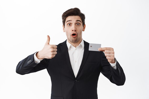 Amazed entrepreneur recommend bank, shows thumbs up and credit card, standing impressed in suit against white background
